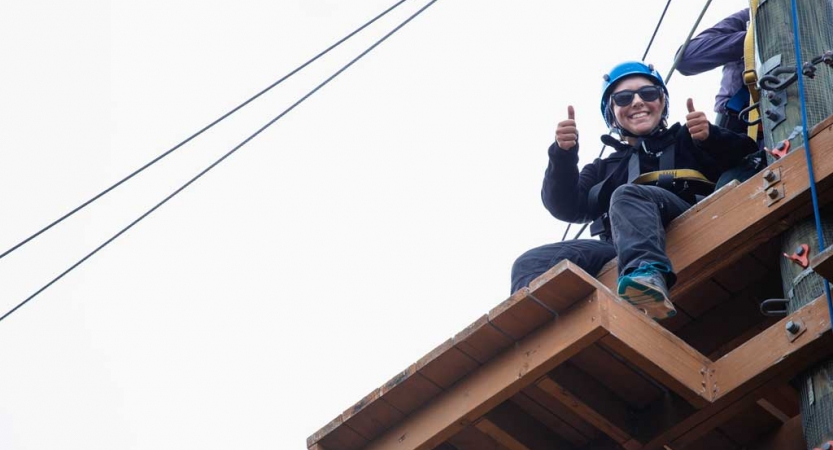 From a ropes course platform, a person wearing safety gear looks down at the camera, smiling, and gives two thumbs up